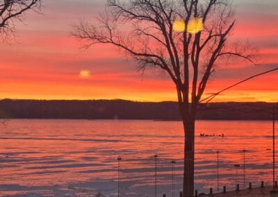 A vibrant sunset over a calm lake with orange, pink, and purple hues in the sky. Red Pines Onalaska stands nearby as bare trees and a wooden dock are reflected in the tranquil water.