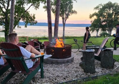 People relax in Adirondack chairs around a fire pit near a lake at sunset at Red Pines Onalaska. Some sit, while others stand or walk nearby. Trees frame the scene, and a bike leans against a tree, creating a peaceful outdoor atmosphere.