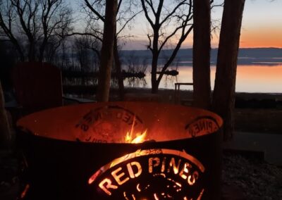 A metal fire pit with "Red Pines Bar & Grill" cut into it glows with flames, set outdoors near leafless trees by a calm lake at sunset—capturing the cozy atmosphere at Red Pines Onalaska.