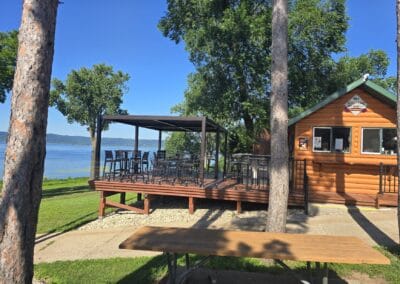 A wooden lakeside cabin at Red Pines Onalaska features a shaded deck with tables and chairs overlooking the water; tall trees and green grass surround the area, and a picnic table sits in the foreground under sunlight.