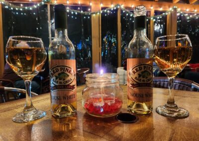 Two glasses and two bottles of white wine sit on a wooden table at Red Pines Onalaska, with a lit candle in the center. Twinkling string lights and blurred outdoor scenery create a cozy, festive atmosphere in the background.
