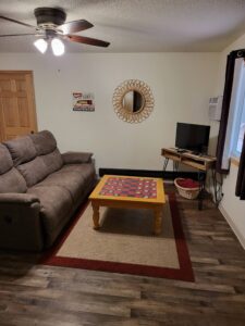 A cozy living room at Red Pines Onalaska features a brown recliner sofa, wooden coffee table with a checkerboard, small TV on a stand, round decorative mirror, wall art, and a window with dark curtains.
