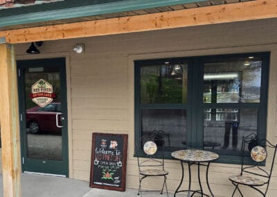 A small outdoor seating area with two chairs and a round mosaic table sits in front of the Red Pines Onalaska lodge entrance. A chalkboard sign welcomes visitors, and a glass door displays the lodge’s name and logo.