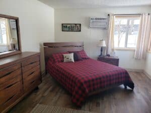 A cozy bedroom at Red Pines Onalaska with a wooden bed and dresser, red and black plaid bedding, a bedside table with a lamp, beige curtains over the window, and an air conditioner above. Snowy views complete the warm retreat.