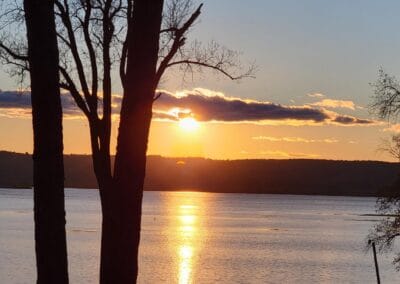 The sun sets over a calm lake at Red Pines Onalaska, casting a golden reflection on the water, with silhouetted trees in the foreground and distant hills under a partly cloudy sky.