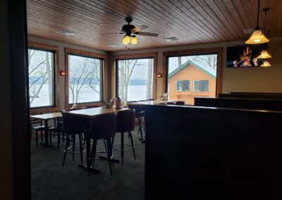 A cozy Red Pines Onalaska restaurant dining area with large windows overlooking a lake and trees. There are wooden tables and chairs, a wooden ceiling, a ceiling fan, and a TV displaying a fireplace on the wall.