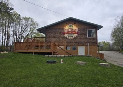 A brown wooden building with a deck and a sign reading "Red Pines On The Lake Lodging & Cafe" sits on a grassy hill by the road, surrounded by trees under a cloudy sky near Red Pines Onalaska.