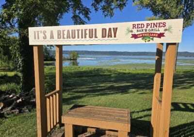 A wooden frame with a sign reads "It's a Beautiful Day" and "Red Pines Bar & Grill on the Lake," set in a grassy area at Red Pines Onalaska, overlooking a lake and distant hills under a clear blue sky.