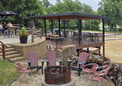 Outdoor seating area with pink Adirondack and metal chairs around a fire pit, next to a covered wooden deck, sandy volleyball court, trees, and people dining on a patio in the background.