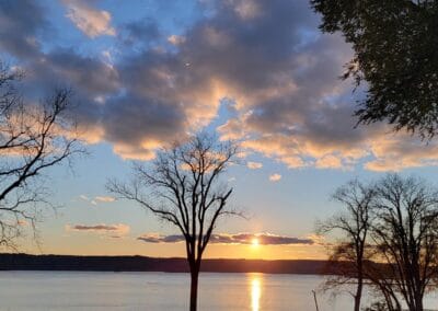 Sunset over a calm Lake Onalaska with the sun reflecting on the water, silhouetted bare trees in the foreground, and clouds scattered across the blue sky.