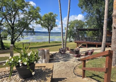 A lakeside patio at Red Pines Onalaska features metal chairs around a fire pit, a shaded deck, and lush green grass. Trees frame views of the calm lake beneath a blue sky. A potted plant rests on a stone ledge in the foreground.