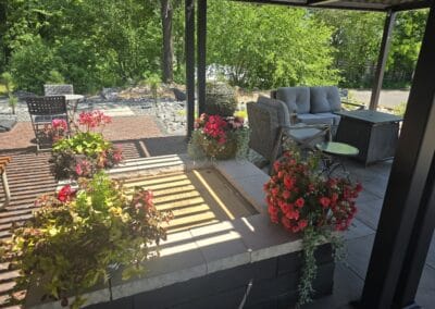 A shaded patio area with cozy seating, potted flowers, a sofa, and chairs under a pergola, surrounded by lush green trees and sunlight casting shadows through the roof slats.