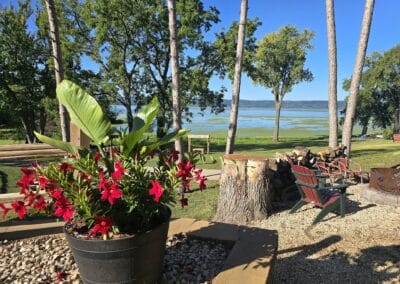 A large black flower pot with vibrant red flowers sits in a landscaped area at Red Pines Onalaska, overlooking green grass, tall trees, wooden chairs, and a scenic view of a lake under a clear blue sky.